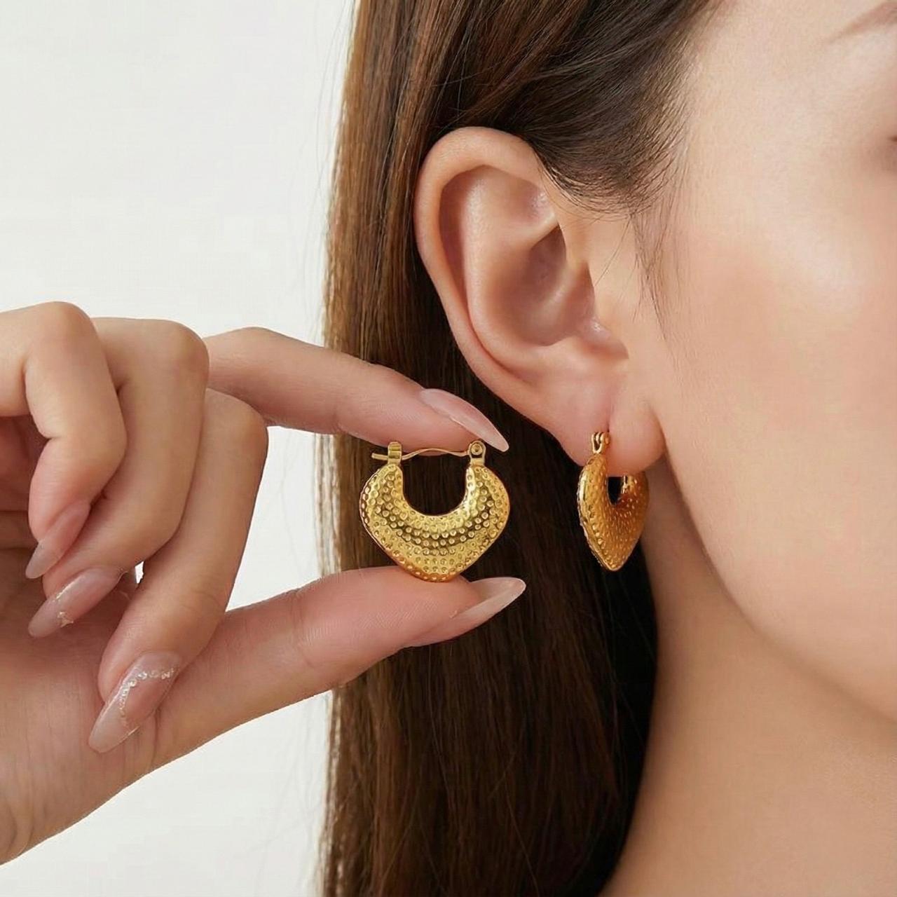 Gold hoop earrings being worn and held by a hand against a neutral background