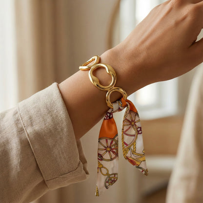 Gold bracelet with a patterned scarf on a wrist against a neutral background